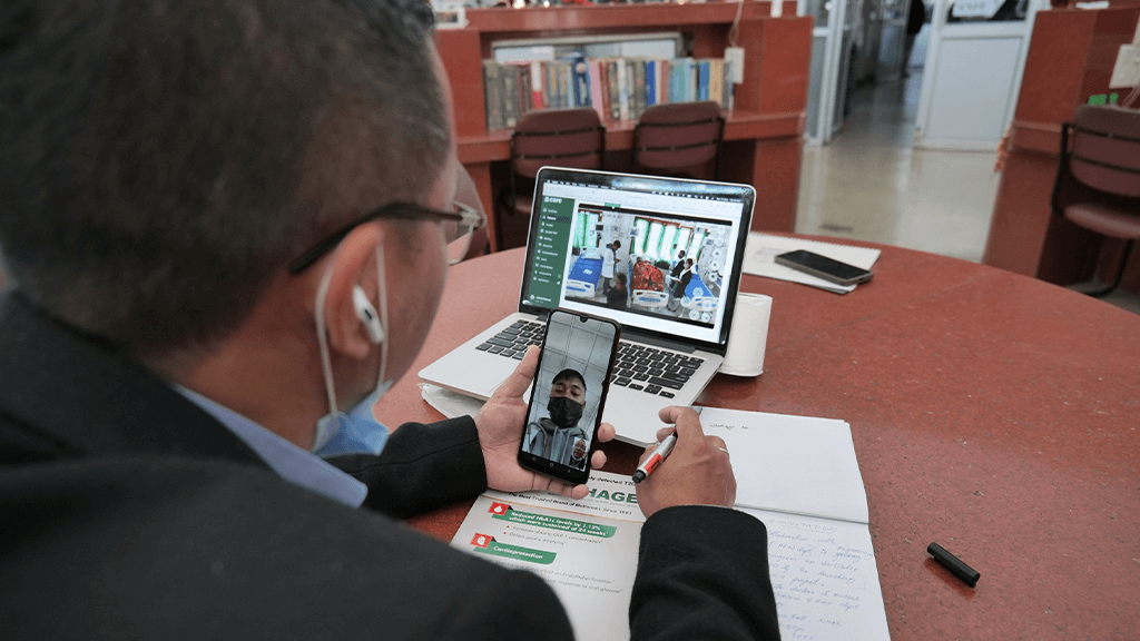 Image of a doctor is on a video call with another doctor on a mobile phone. The doctor is in front of a laptop that is displaying a dashboard of the CARE software. The dashboard shows a live view of a remote ICU.